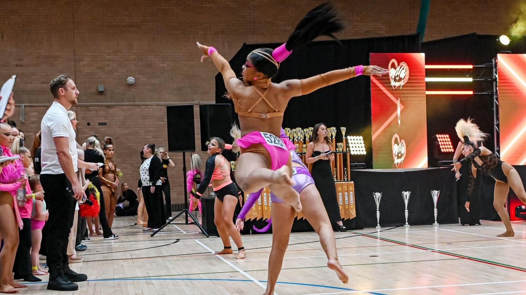 A young woman dances in a carnival-style outfit as part of a dance competition in a large sports hall, while dozens of other dancers watch on.