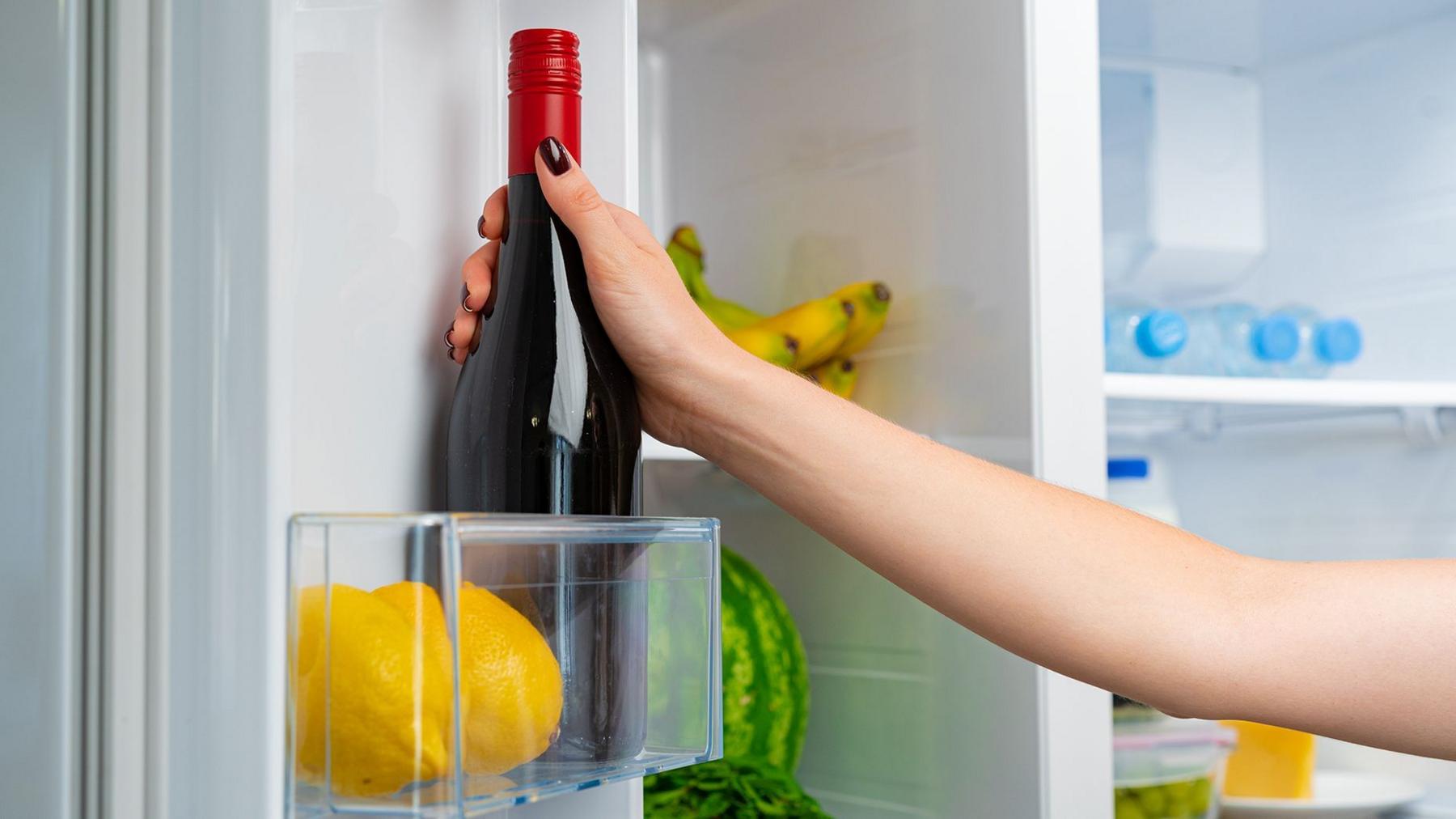 A hand placing a dark glass bottle with a red screw cap into an open refrigerator. In the foreground, two yellow lemons sit in a clear door shelf compartment. Inside the fridge, a watermelon, a bunch of bananas, several plastic water bottles, a lidded food container and a block of cheese are visible on shelves. The refrigerator interior is white and brightly lit.