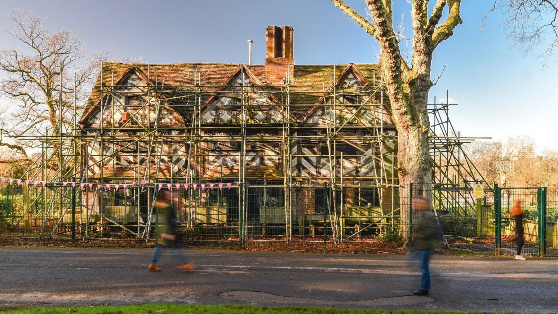 A 16th Century pub with a thatched roof covered with scaffolding. Trees and people can be seen in front of the pub.