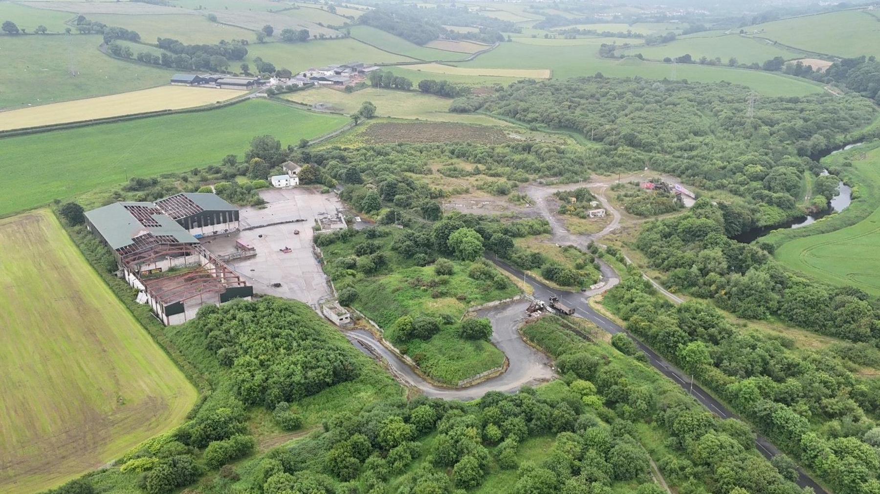 the site of an illegal dump sits surrounded by green fields and countryside. a number of dilapidated buildings are on the left, while minor roads run across the width of the site
