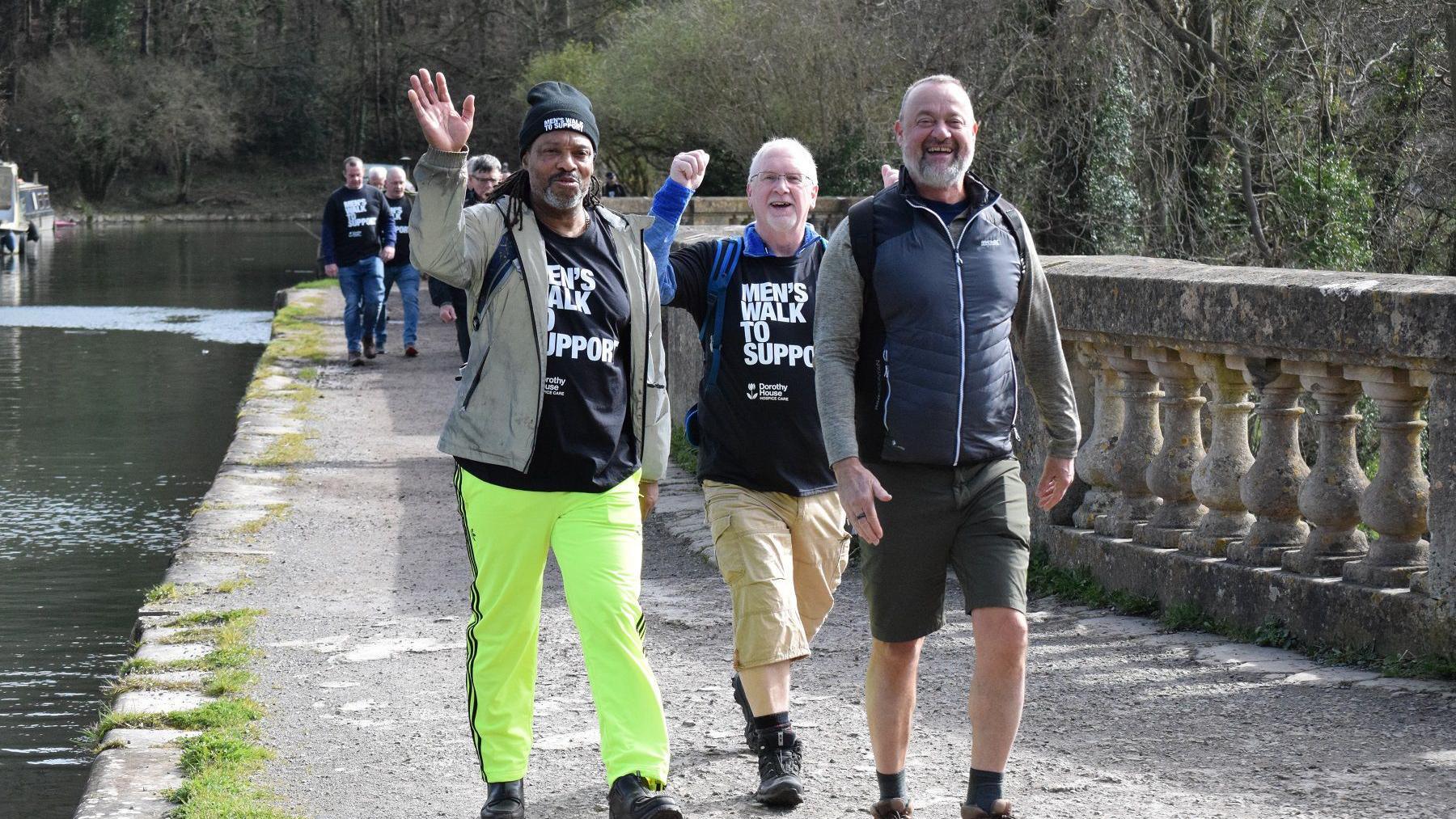 Three men smile at the camera as they walk by a river. Two of them raise their hands in the air and they wear T-shirts which say 'Men's walk to support'. There is a small group of men walking behind them.