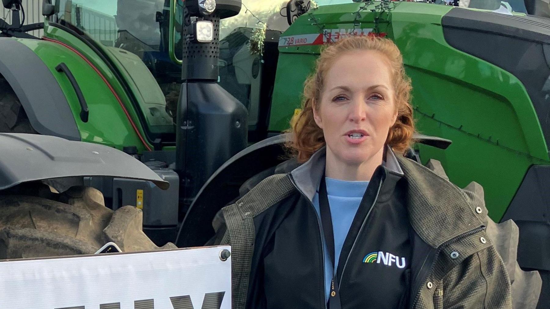 Clare Wise is standing in front of a green tractor and looking directly into the camera. She has short, curly ginger hair and is wearing a jacket with the "NFU" logo on it.