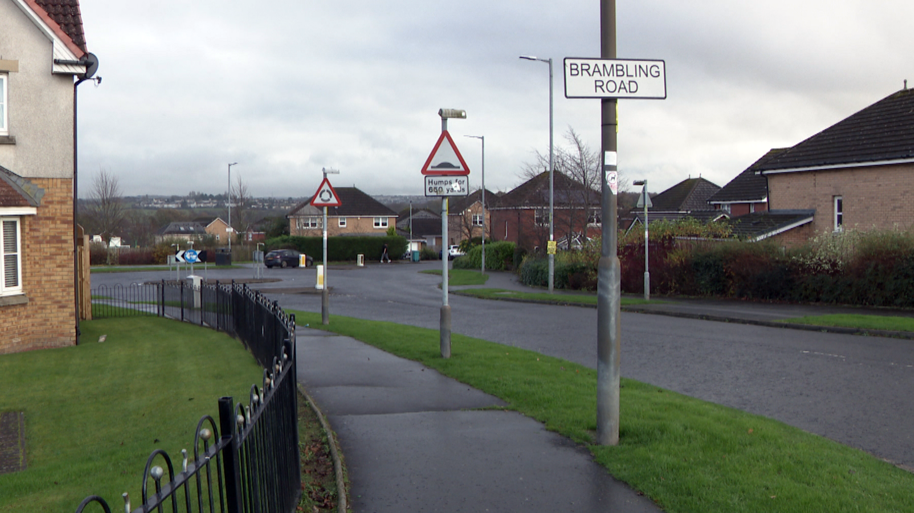 Road approaching a roundabout with modern houses on either side. There is a sign saying Brambling Road as well as one depicting speed humps and another a roundabout.