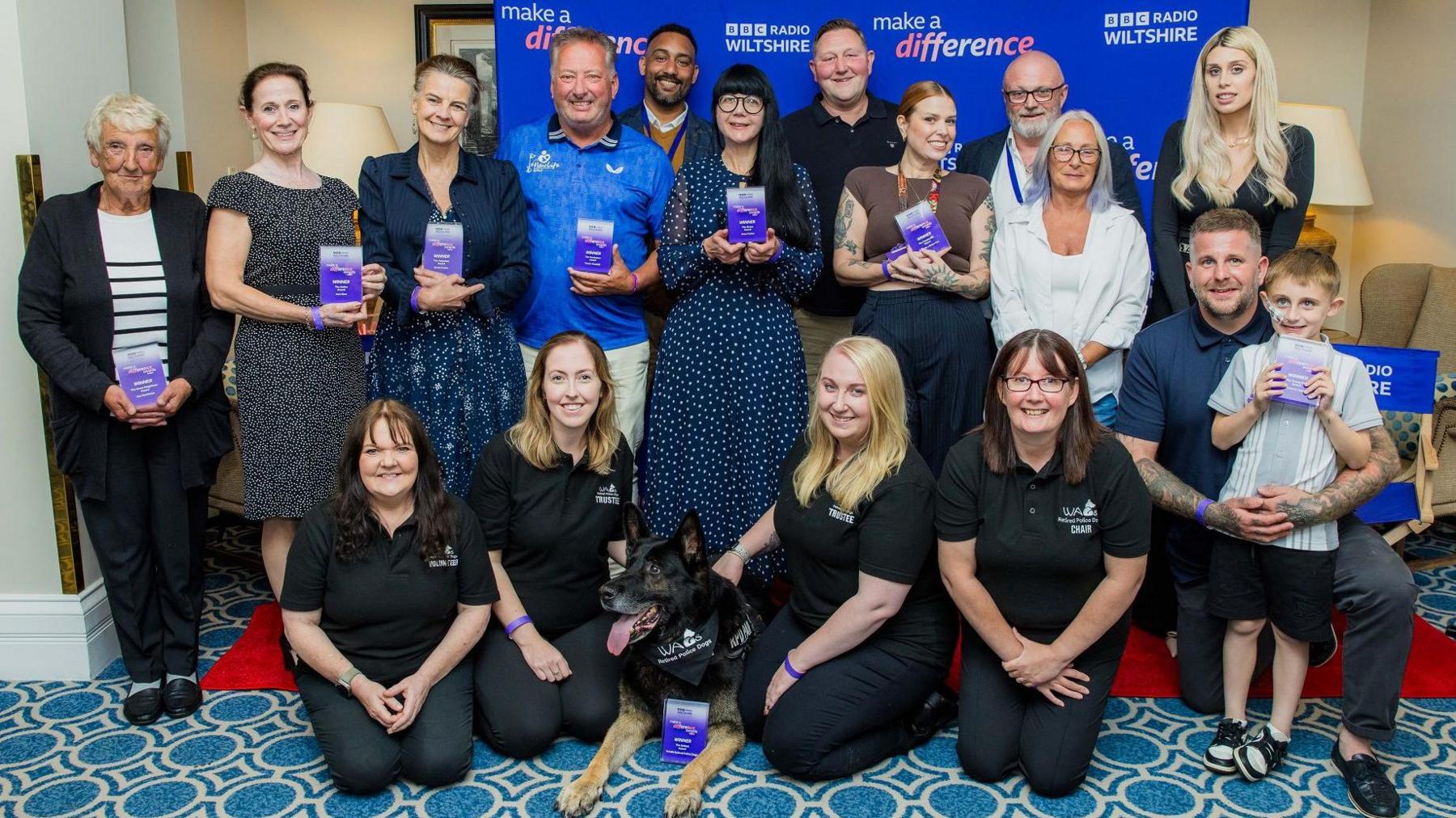 A group of people - standing and kneeling on the floor. All smiling at the camera standing in front of a purple BBC Radio Wiltshire banner. Many are holding small purple glass awards