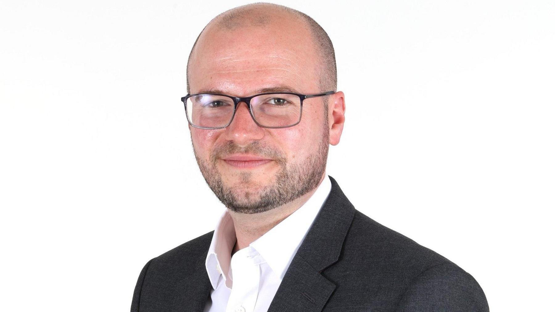 A corporate headshot of a man in a dark suit and open-necked shirt. He has a close-shaven head and short beard, and is wearing glasses.