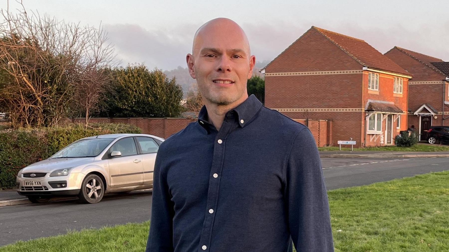 A photo showing a white man, he is bald and is wearing a blue shirt with white buttons. He is stood on a green stretch of grass, in the background is a silver car and a block of red brick homes