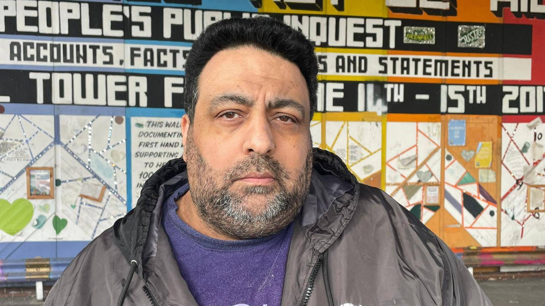 Nibil Choucair is seen with short black hair and a beard stands in front of a colorful mural about the Grenfell Tower fire, wearing a black jacket and a purple T-shirt.