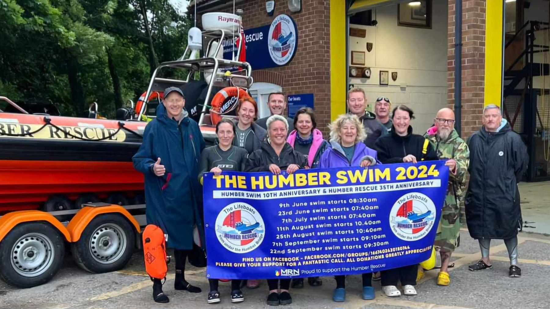 Twelve men and women standing in front of a Humber Rescue boat holding a big blue sign with yellow and white writing. The sign reads "The Humber Swim 2024" and has a list of dates of when the swims are taking place below.