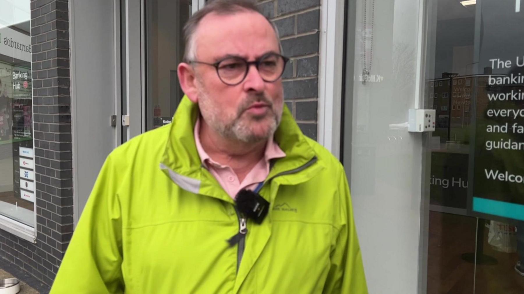 Phil Dunn is outside the banking hub. He is wearing a green hi-vis jacket and has round glasses. 