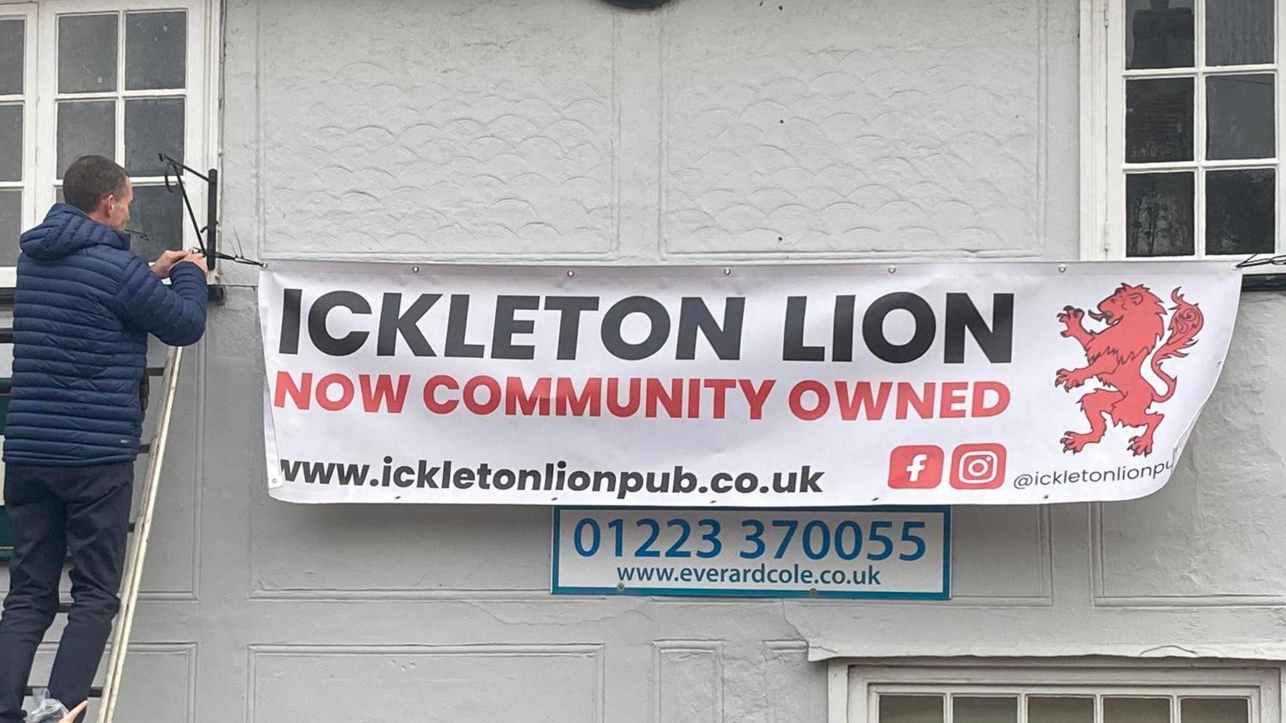 A man is standing on a ladder, against the Ickleton Lion pub, while hanging up a sign. It reads 'Ickleton Lion, now community owned'. 