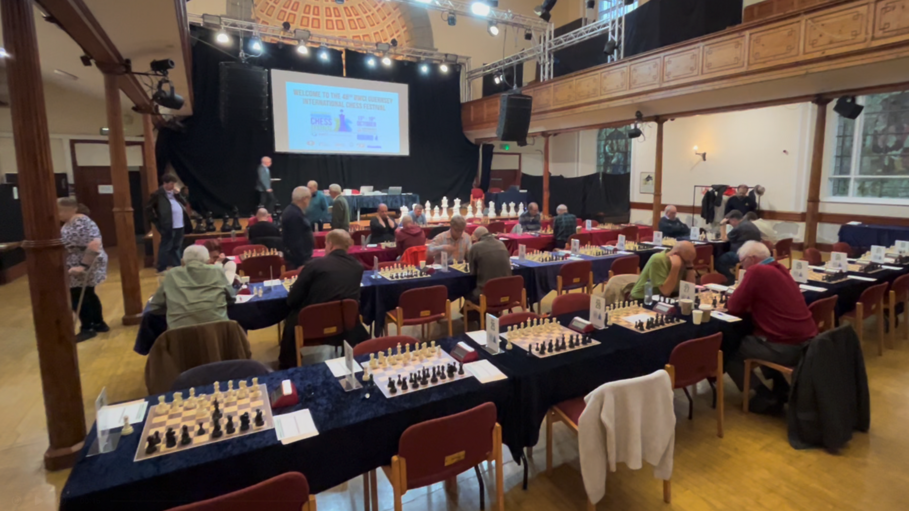 A large performance hall with wooden floors, pillars and balcony, filled with tables and chairs topped by chess sets. It is about half full of people playing chess. A projector on the stage sits behind the judges' table,