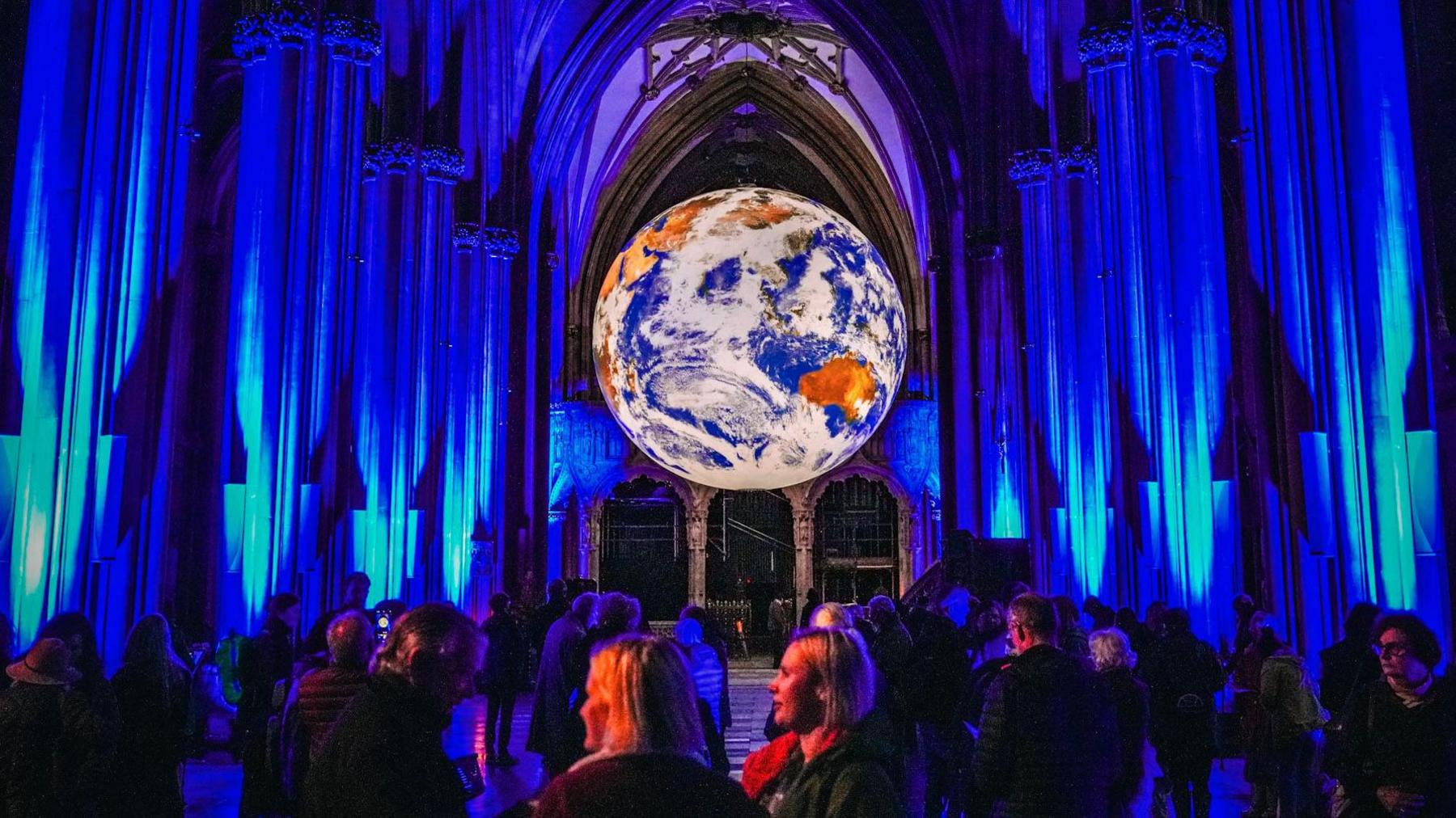 Dozens of people stand under the illuminated Luke Jerram artwork "Gaia" which is a large scale model of the planet Earth. It is hanging from the ceiling in Bristol Cathedral and much of the internal structure of the building is illuminated blue