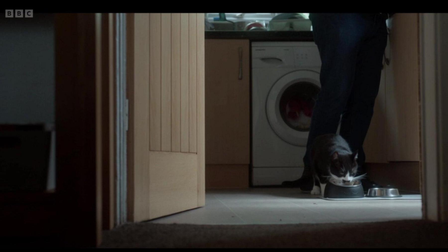 A black and white cat, Spud, eats from a metal food bowl on the floor in a kitchen. Someone's legs wearing black pants are behind him. A washing machine and kitchen cupboards can also be seen behind the cat.