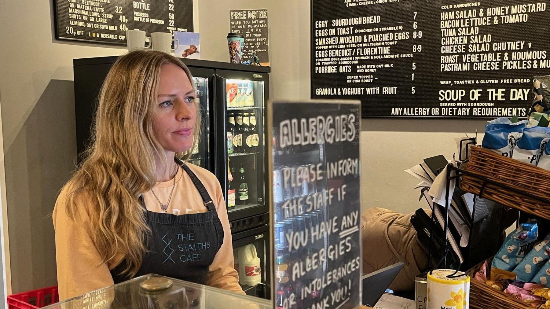 Suzanne Hutton serving behind the counter at the Staiths Cafe. She is wearing a black apron, with the cafe's name written in white, and an orange top. The cafe's menu is displayed on a blackboard to the right of the image with a cooler holding milk and other drinks also in the background. On the counter-top is a sign asking customers to inform staff if they have any food allergies.