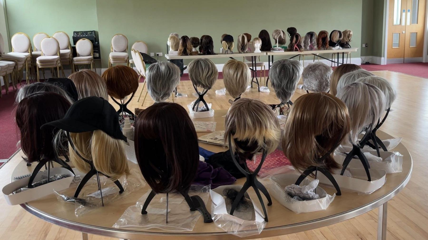 A community hall with two tables filled with different colour and style wigs on stands.
