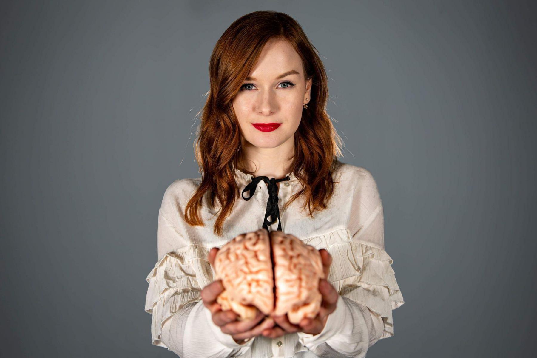 Alice Gray holds up a model of a brain and smiles into the camera. She has long red hair and is wearing a ruffled white shirt.