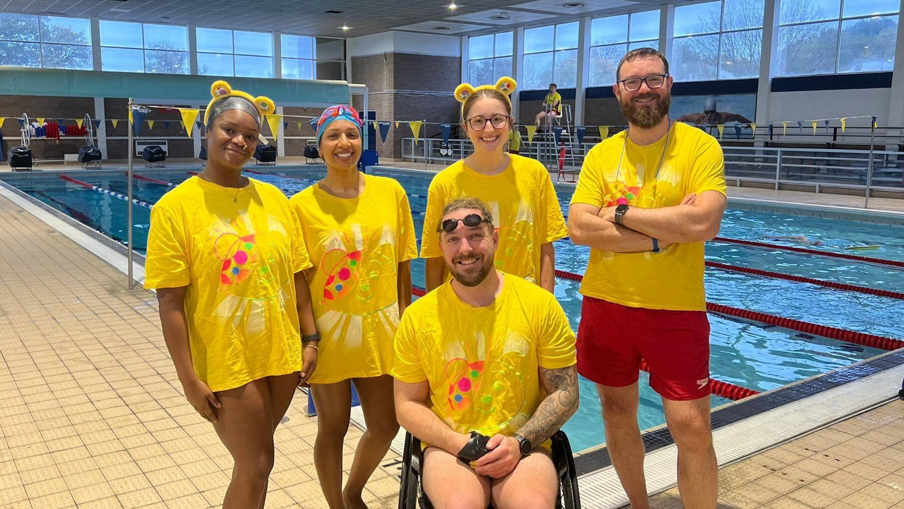 The team of swimmers all wearing yellow children in need shirts. Four of them are stood behind one of them in a wheelchair. Behind them is a swimming pool