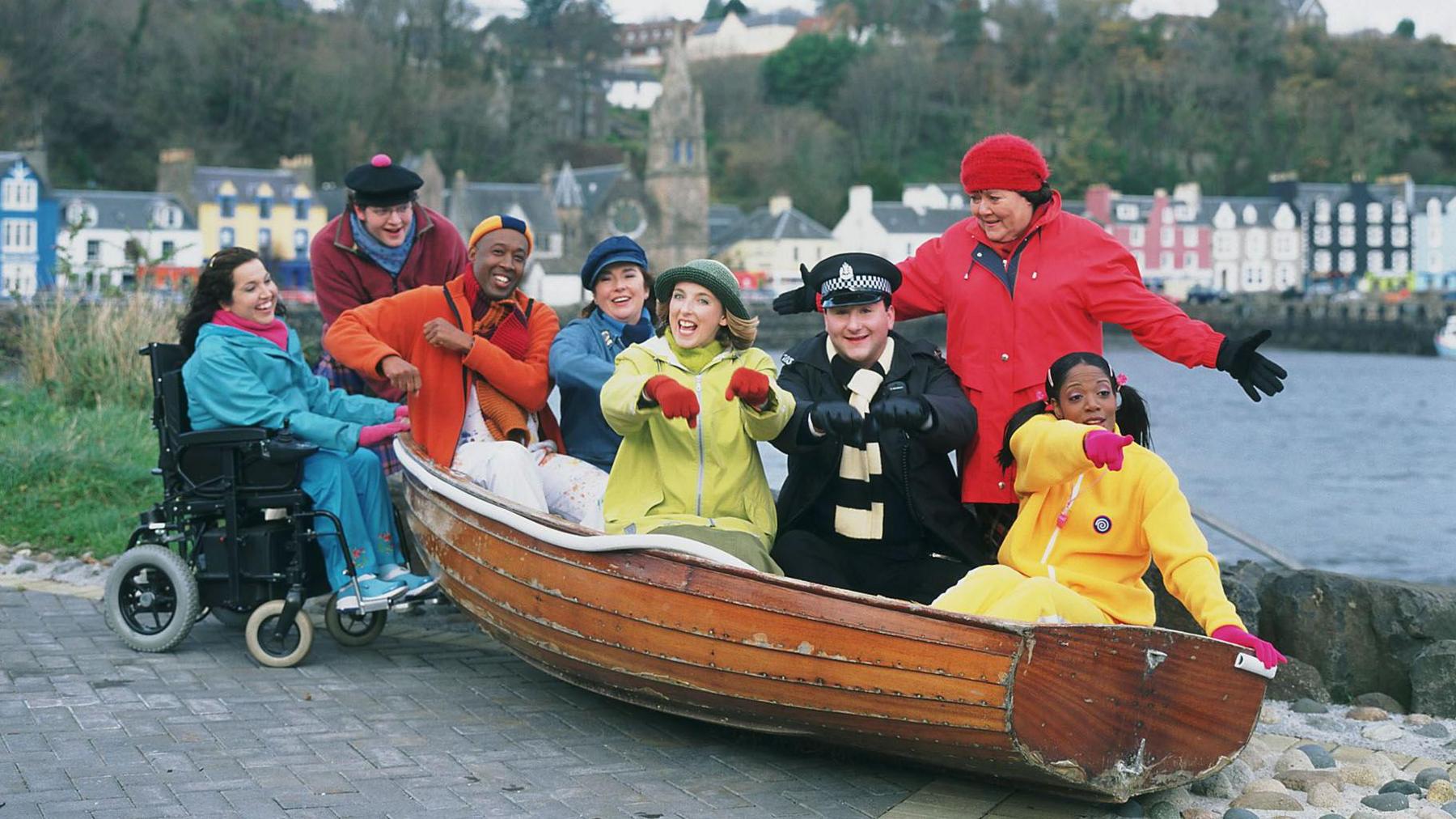 The cast of the original Balamory pose with a wooden rowing boat across the bay from the colourful facades of the houses in Tobermory on Mull