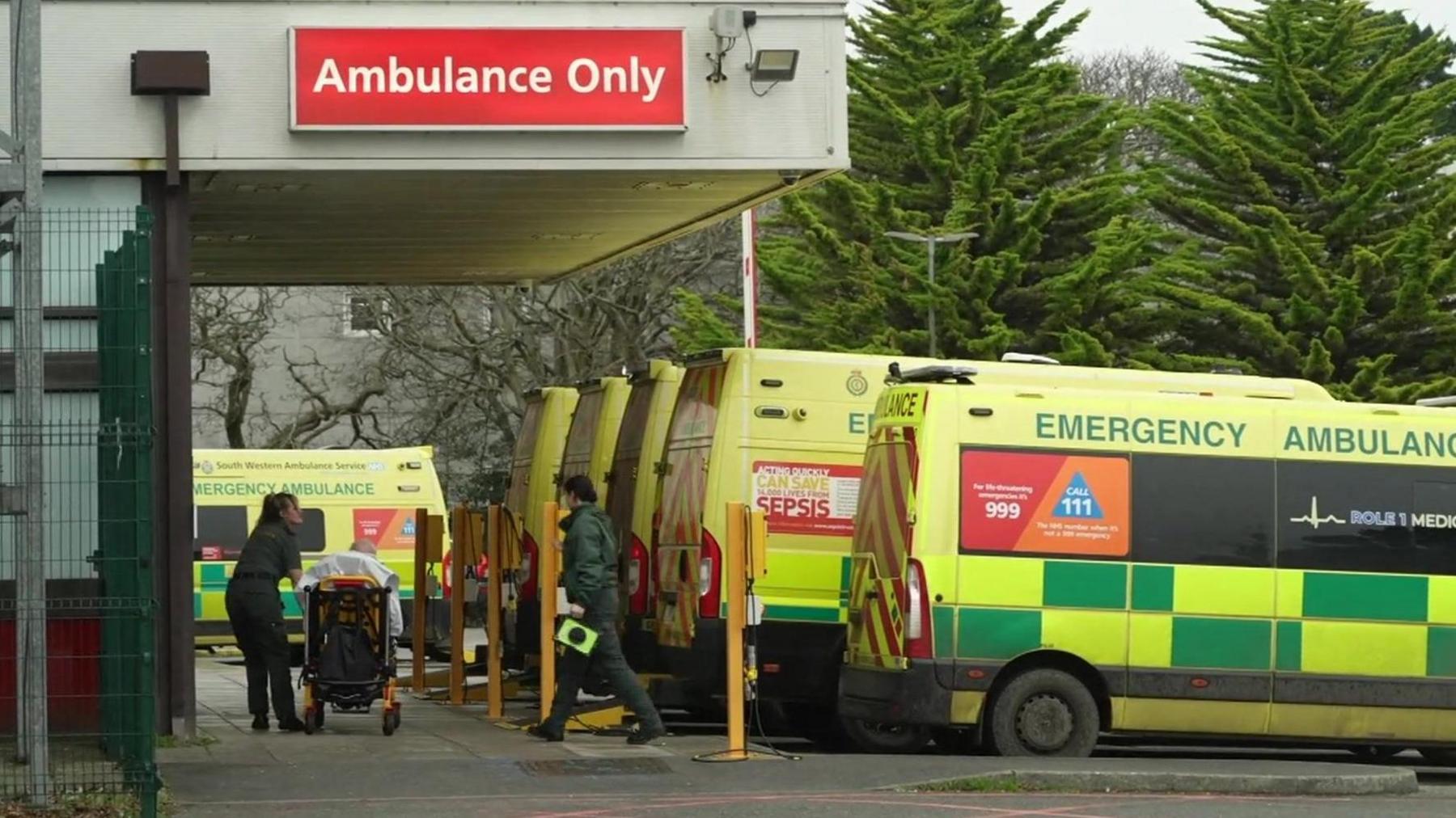 Two paramedics walk away from an ambulance out the Royal Cornwall Hospital in Truro. They are pushing a patient on a bed into the hospital. A sign is on the building which says "ambulance only". There are six ambulances parked up outside the hospital.