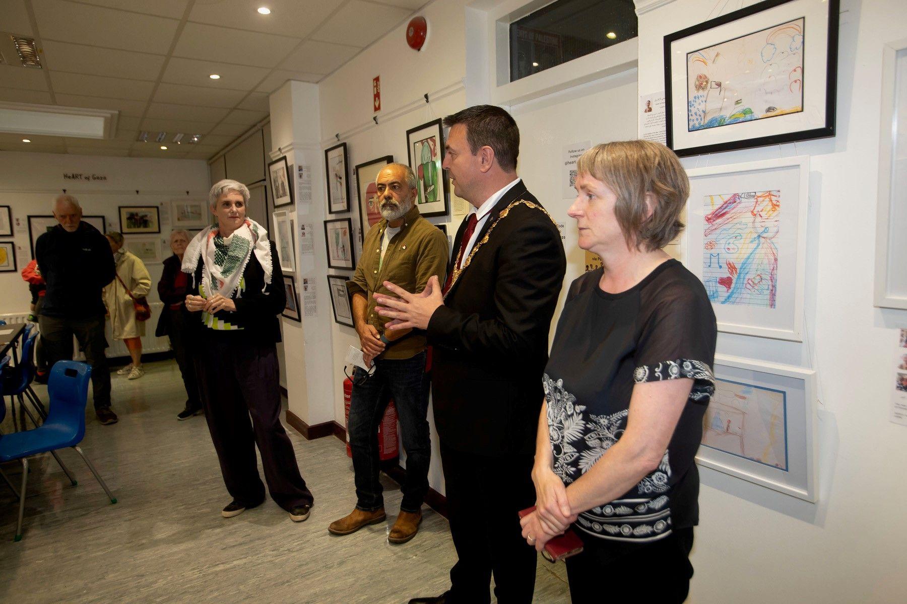 Ruairi McHugh wears a black suit and his gold mayoral chain. He is talking to a group of people in a gallery which has art work on the walls. 