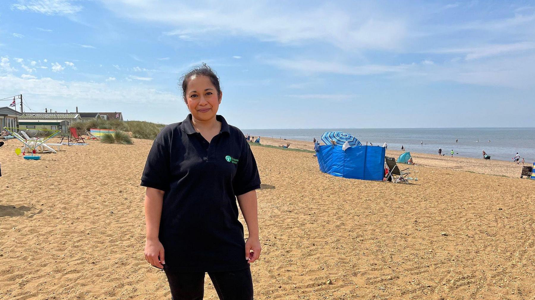 Woman wearing a black t shirt and trousers standing on a coarse sandy beach on a lovely sunny day. Over her right sholder are some beachside properties and her left someone has set up a blue wind break with a striped blue sun umbrella