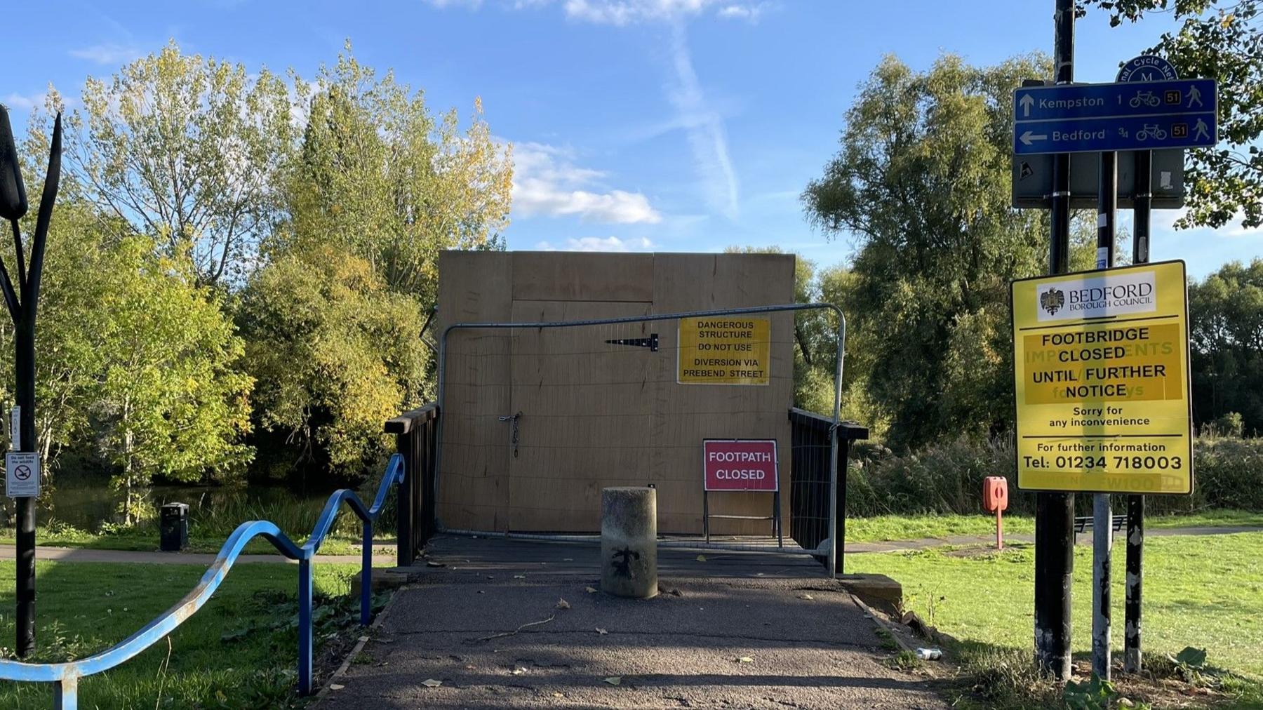 One end of a footbridge which has been boarded up with signs saying "footpath closed". There is a bollard in front of the wooden hoarding.