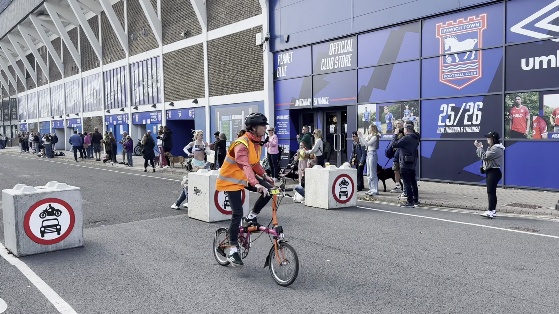 A cyclist wearing high visibility jacket is riding ahead of Holly Archer as she runs down the road with people cheering on the pavement