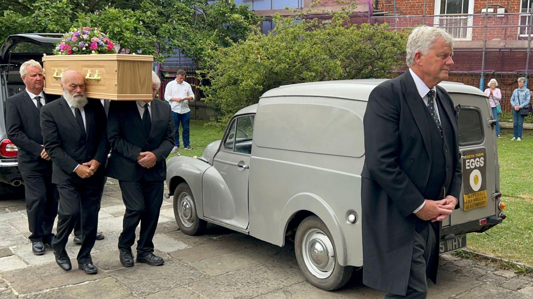 Four men carrying the casket of Wilf Peddle on their shoulders as they walk into a church, led by the foreman. The morris minor that Wilf Peddle would drive to deliver eggs can be seen parked in the background