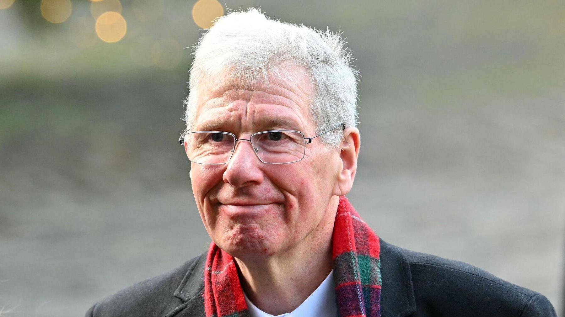 Kenny MacAskill smiles off to the side. He has short white hair and wears silver glasses. He wears a dark coat and white shirt and has a tartan scarf around his neck.