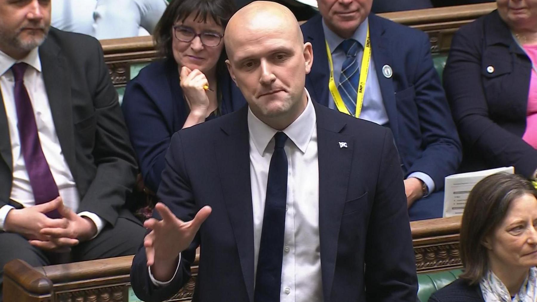 Stephen Flynn speaking from the green SNP benches in the House of Commons. He has a bald head and is wearing a blue suit jacket, a dark tie and a white shirt. He is gesticulating with his hand and there are other politicians around him. 