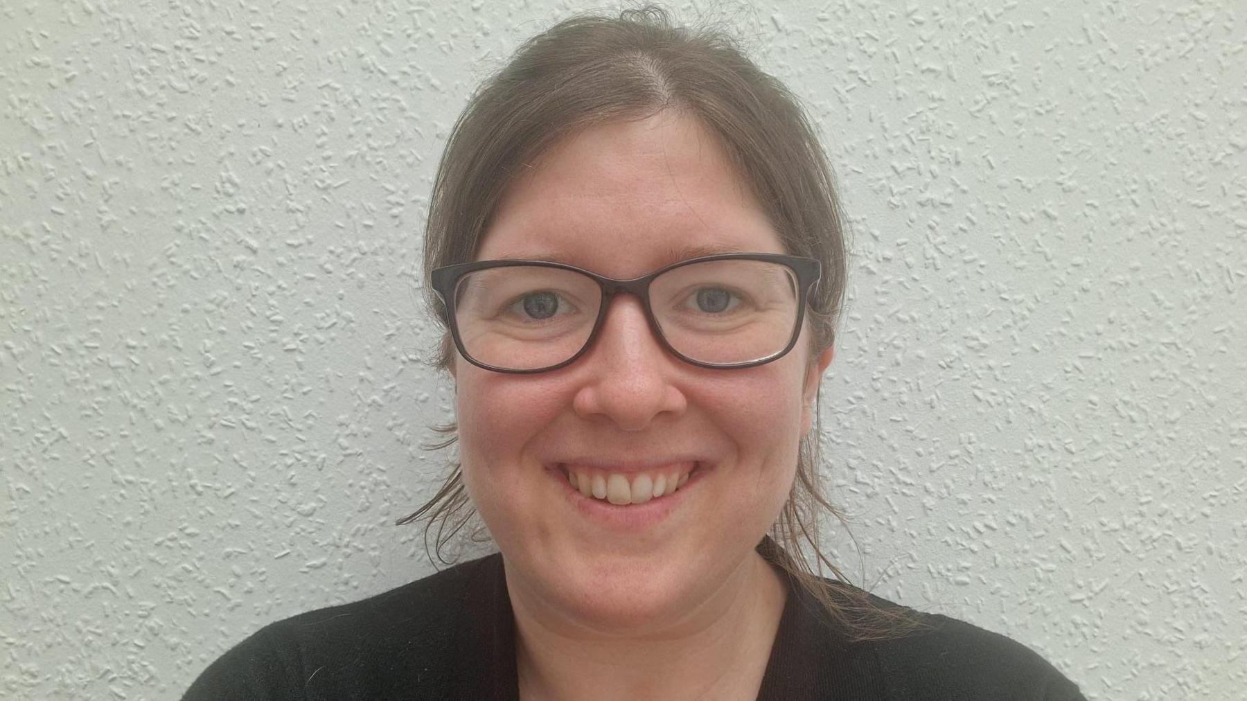 Emily smiles at the camera against a plain background. She is wearing a black top and has brown hair tucked behind her ears. She is also wearing black square-framed glasses.