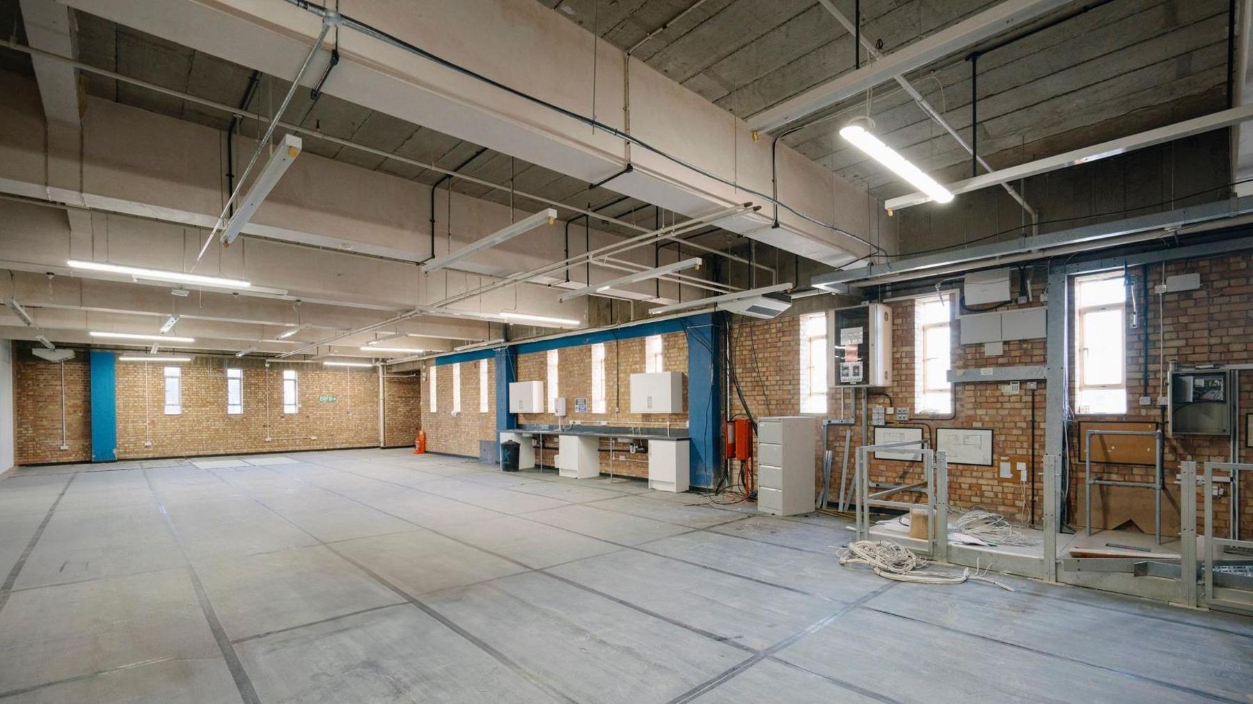 An empty warehouse with small windows. The room has some tools in it, with the walls lightly coloured brick.