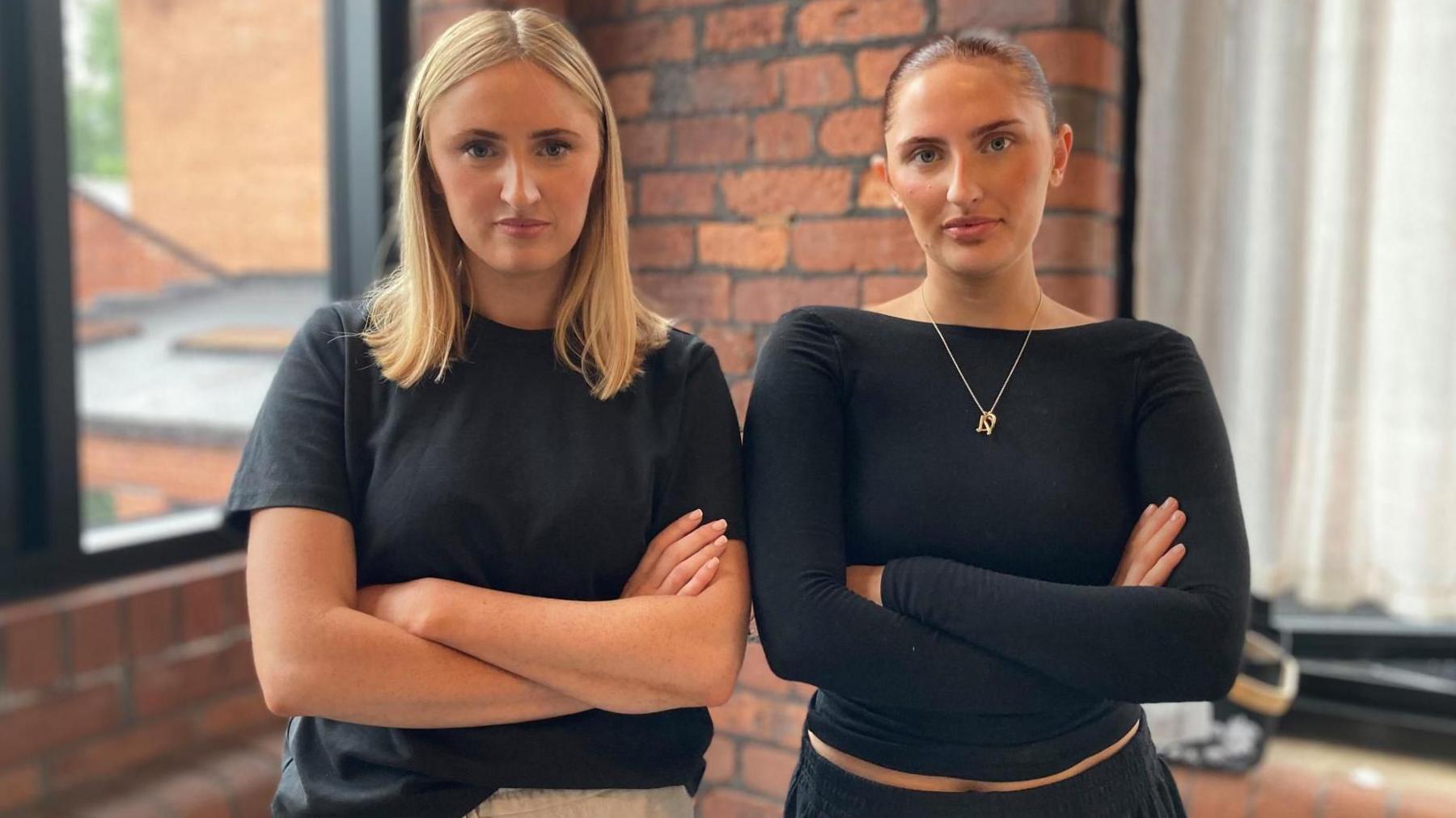 Two young women stand with folded arms looking angry