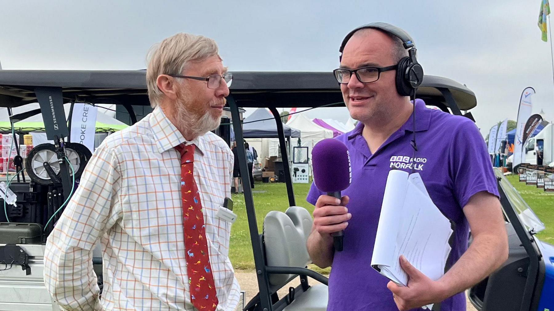 Chris Goreham on the right is standing outside in a field in front of a golf buggy. He is wearing a purple branded BBC Radio Norfolk T-shirt and is holding a purple microphone and clipboard. He is standing next to a man, who is on the left, and he is wearing a chequered white shirt and red tie. They are standing at the Norfolk Showground at the Royal Norfolk Show.