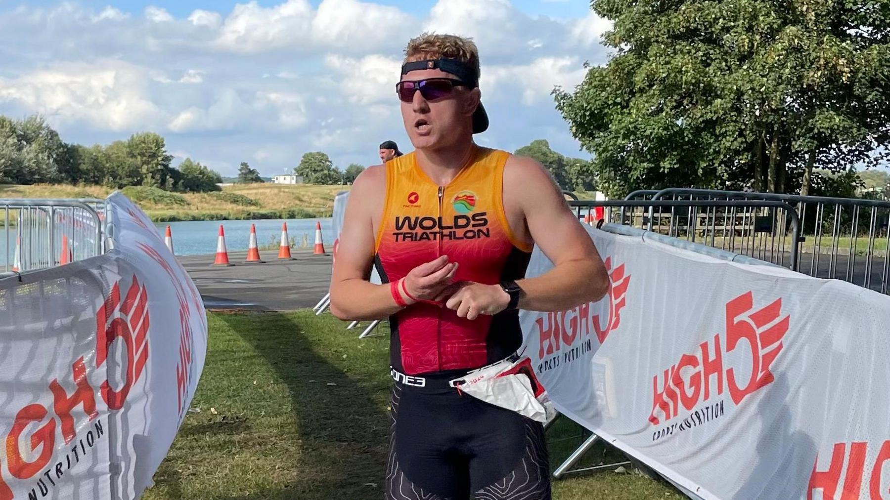 Angus runs towards what looks like the finish line of a triathlon - there is a lake in the background with cones and metal barracades funneling him through onto a grass area. He wears a red lycra top with Wolds triathlon written on.