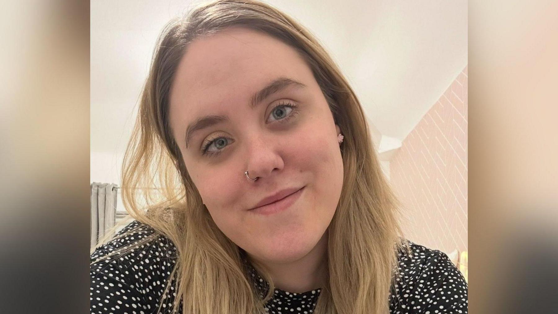 A close-up picture of a smiling woman with long blonde hair. She is wearing a nose ring and a black and white spotted dress.