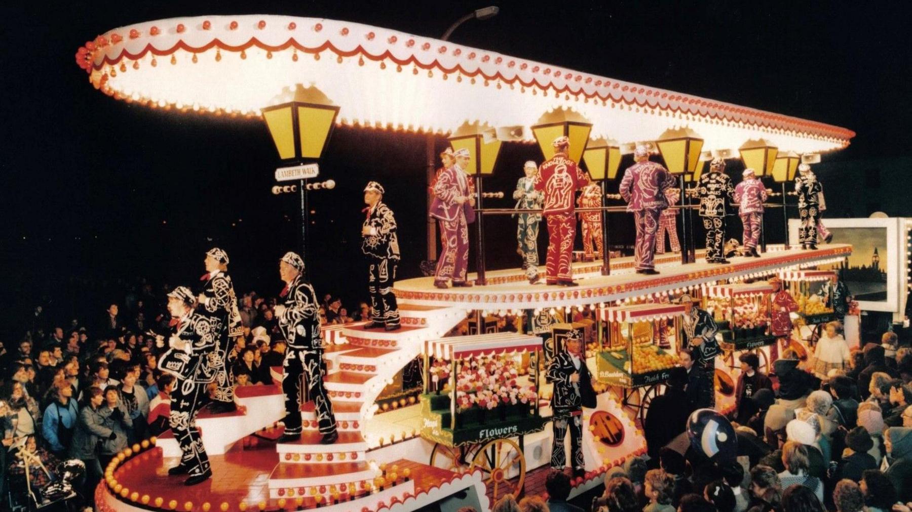 A lit up carnival cart during a procession in 1985. Dancers are standing on the cart, dressed in colourful suits. There is a big crowd watching.