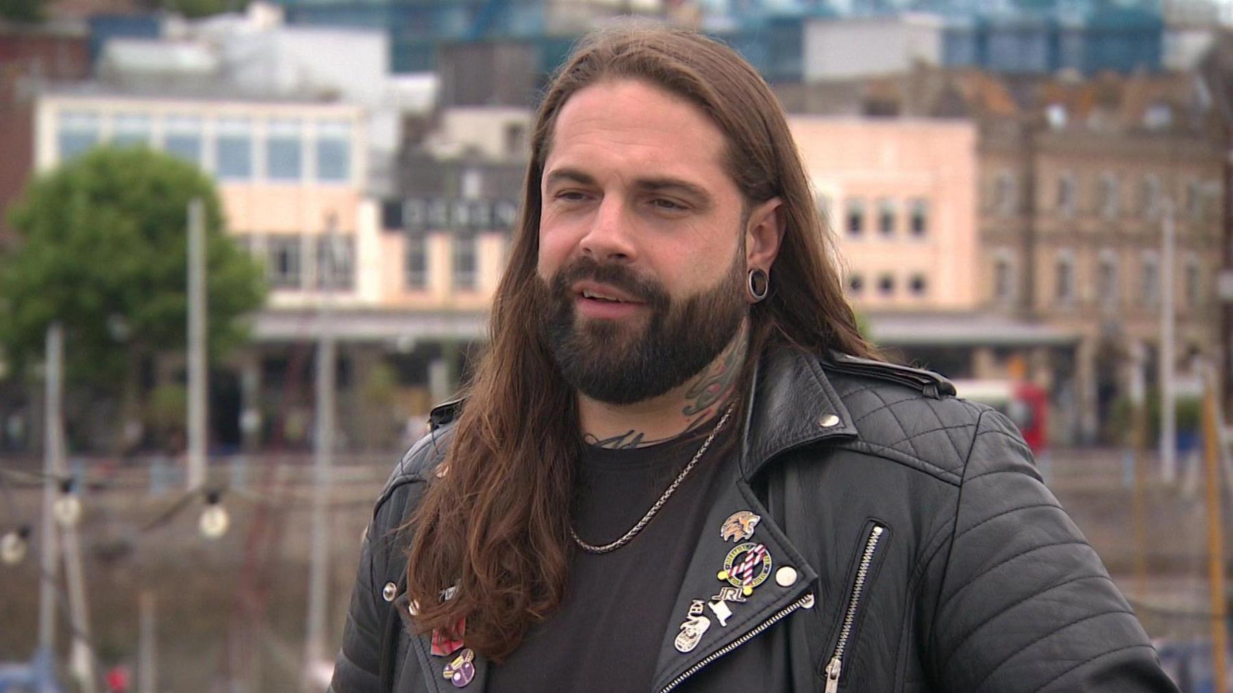 A man with long brown hair and a dark beard stood in front of a harbour and some buildings. He is wearing a black leather jacket which has various pins and badges on the lapel.