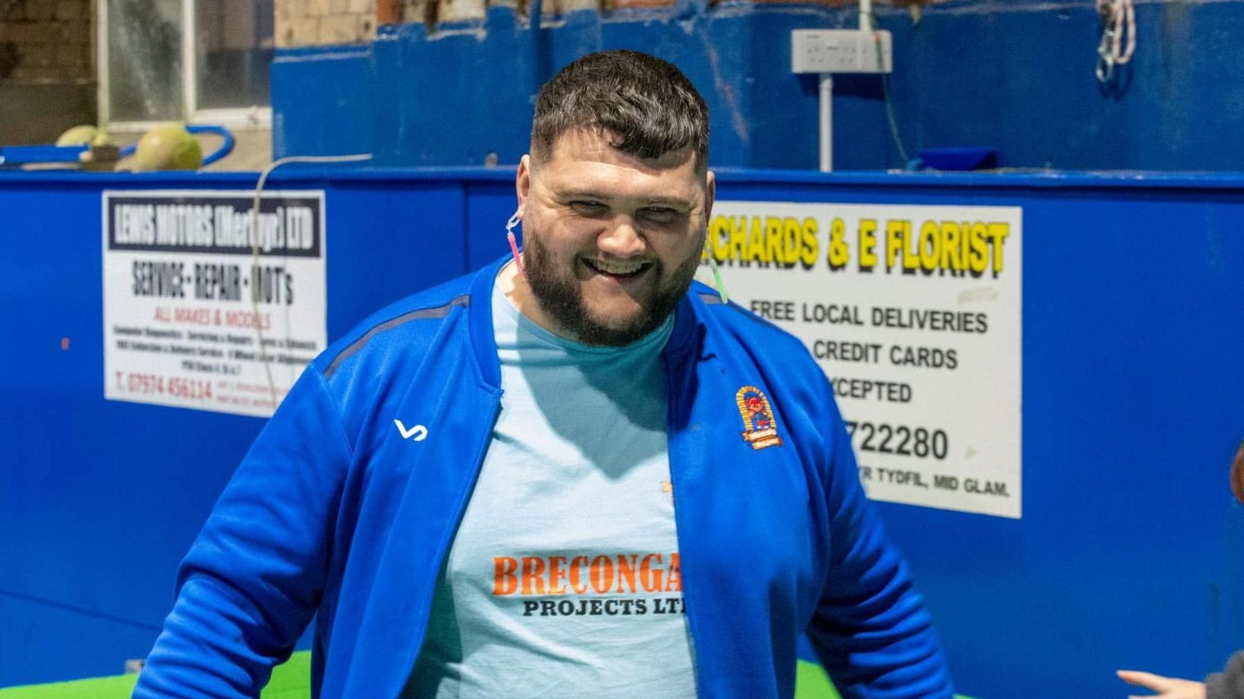 Luke Palmer smiling while coaching at the Diversity Dragons group. He is wearing a grey t-shirt and a blue zip up jacket with the Diversity Dragons logo on the right side.