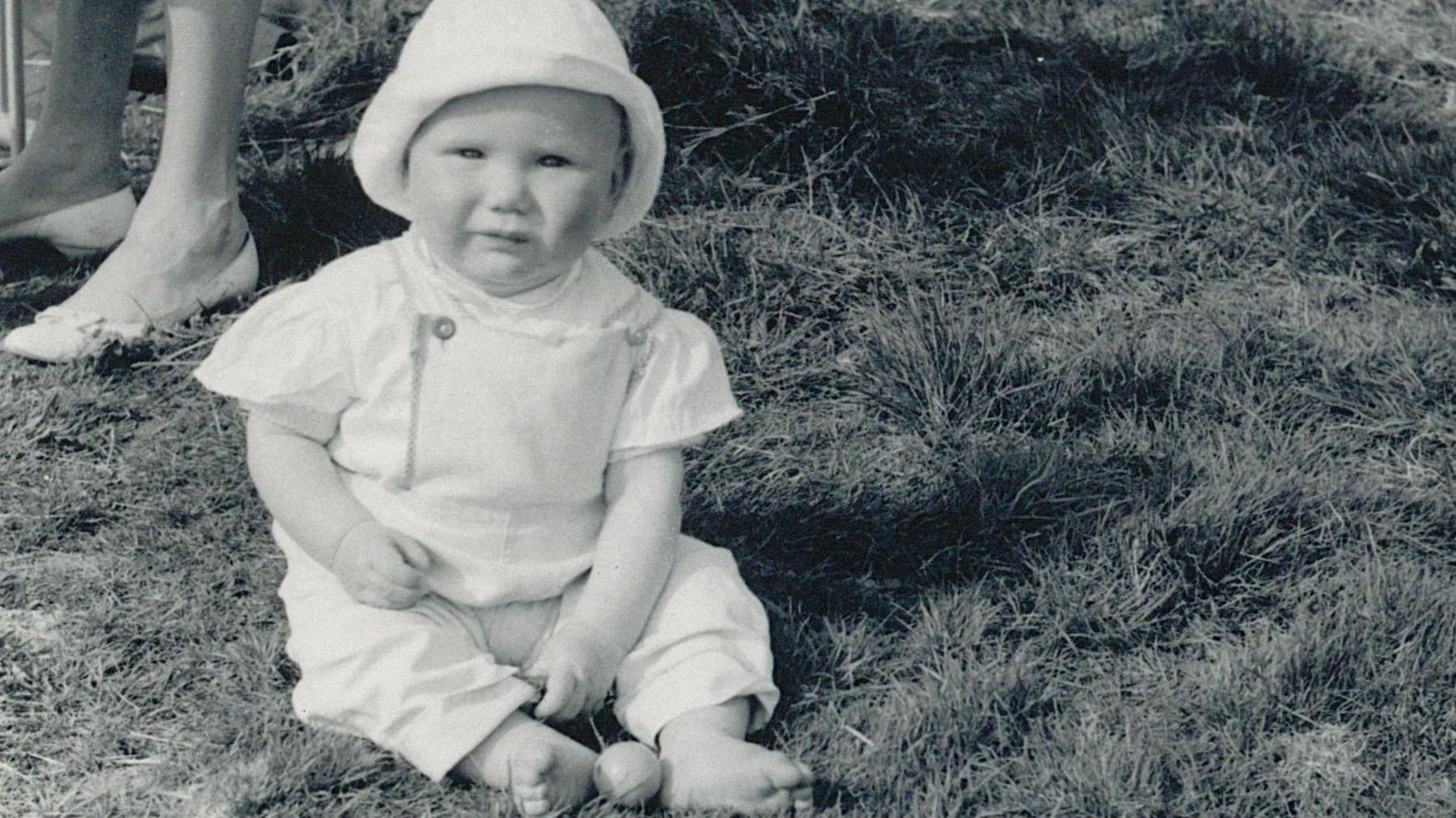 A baby Keir Starmer sitting on grass, dressed in a bonnet and romper suit