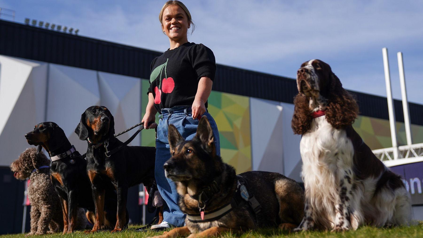 Ellie Simmonds, in black T-shirt and blue denim jeans, stands holding the leads of several dogs on a grass lawn. behind her is the wall of a building. There are five dogs next to her, three sit, one stands and the other lies on the ground.