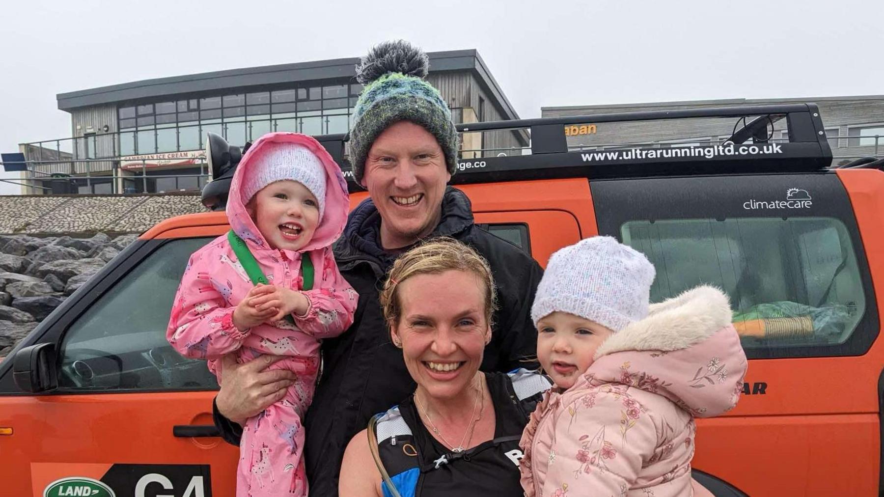 Alison crouches down holding her daughter Seren on her hip. Alison has a watersports vest on and is wearing her hair in plaits. Seren, 3, is wearing a white hat and a pink floral coat with faux fur around the hood. Alison's husband Paul Robinson stands behind them wearing a blue and green hat and a black coat. He is holding daughter Rhian, 5, who is wearinga matching white hat with Seren, and a pink horse cartoon waterproof jumpsuit. The family are on a beach.