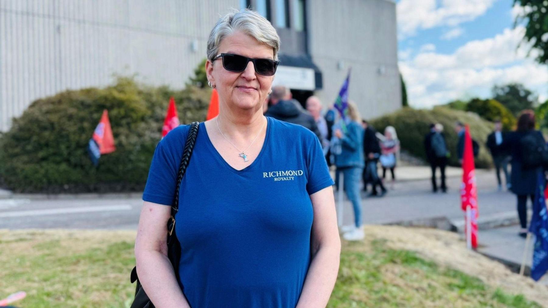 Woman - Charlene Anderson Coltherd - wearing a blue t-shirt and looking a camera, with a council building in the background.