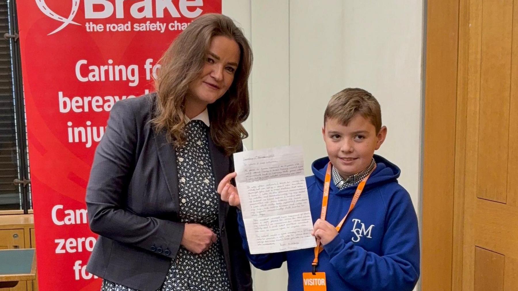 Jenny Riddell-Carpenter stands alongside Roman inside a room. Roman holds his hand written letter on a piece of paper up as he smiles while looking away from the camera. Jenny smiles next to Roman.