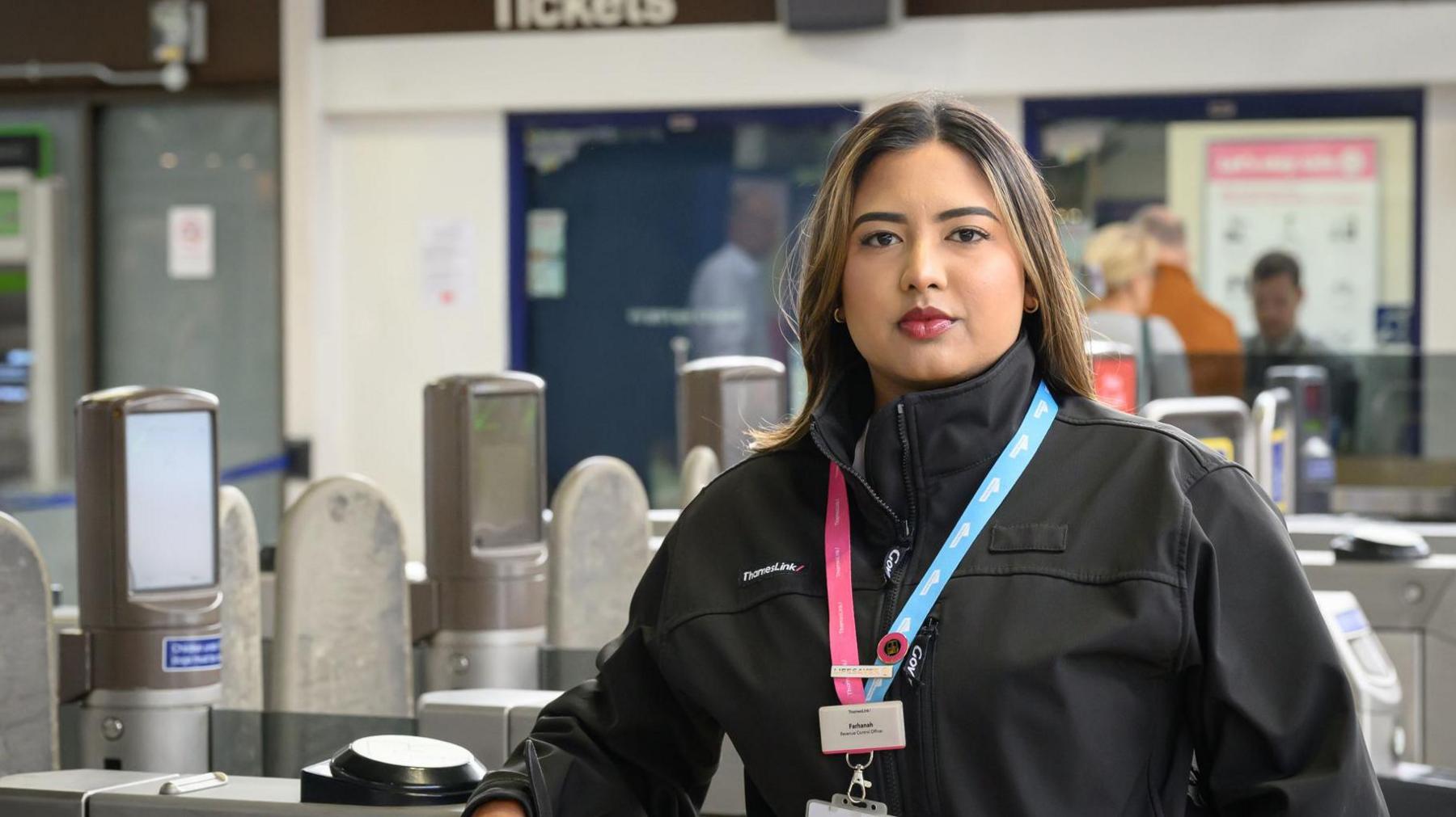 A woman with long brown hair wearing a black top with a pink and blue lanyard stands behind ticket barriers at a train station
