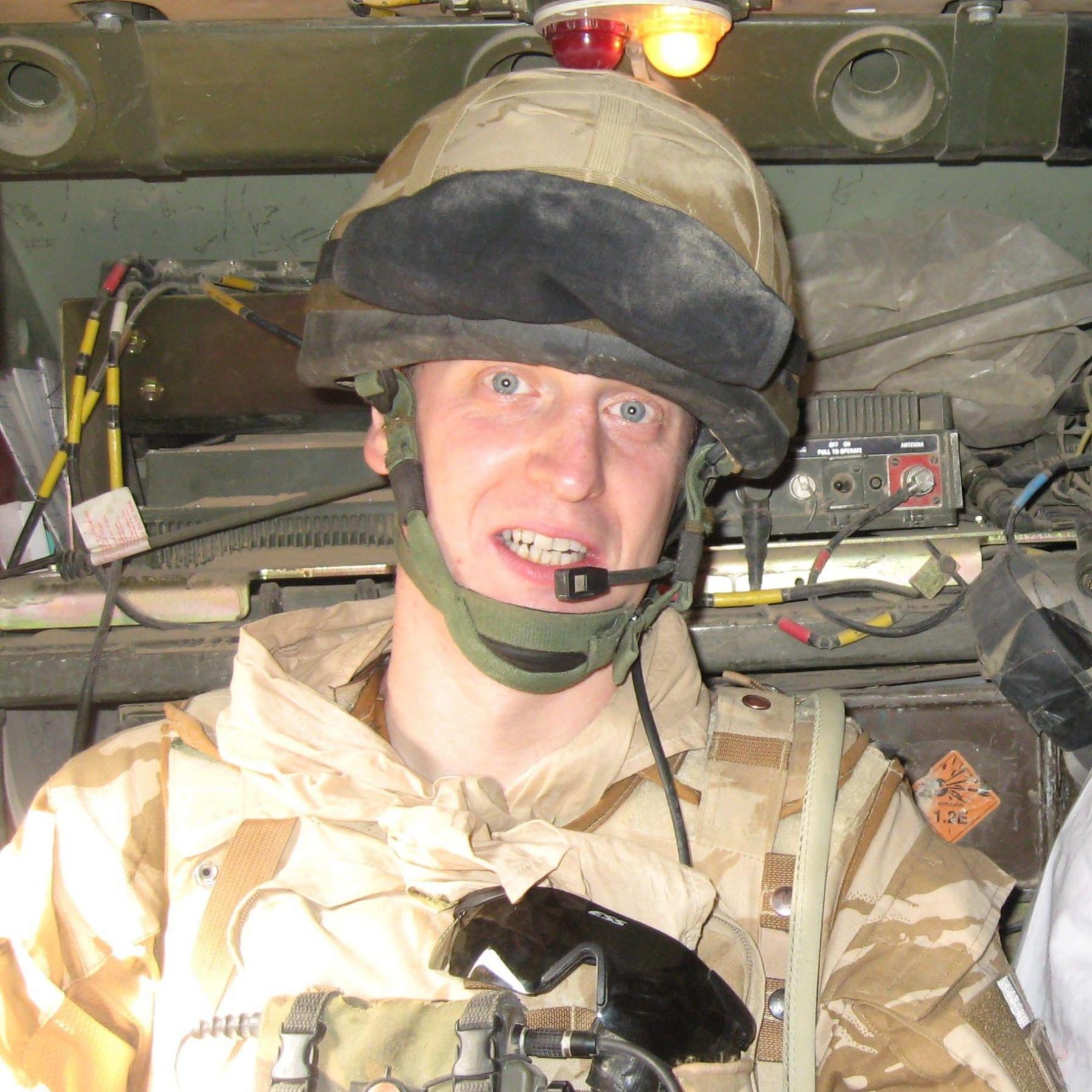 A man dressed in army clothing and a helmet smiles while inside a military vehicle.