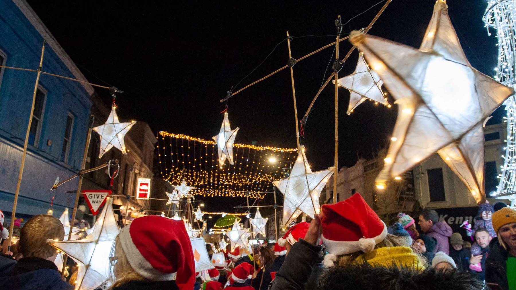 A large group of people wearing red and white Santa style hats. They are standing under giant star lights.