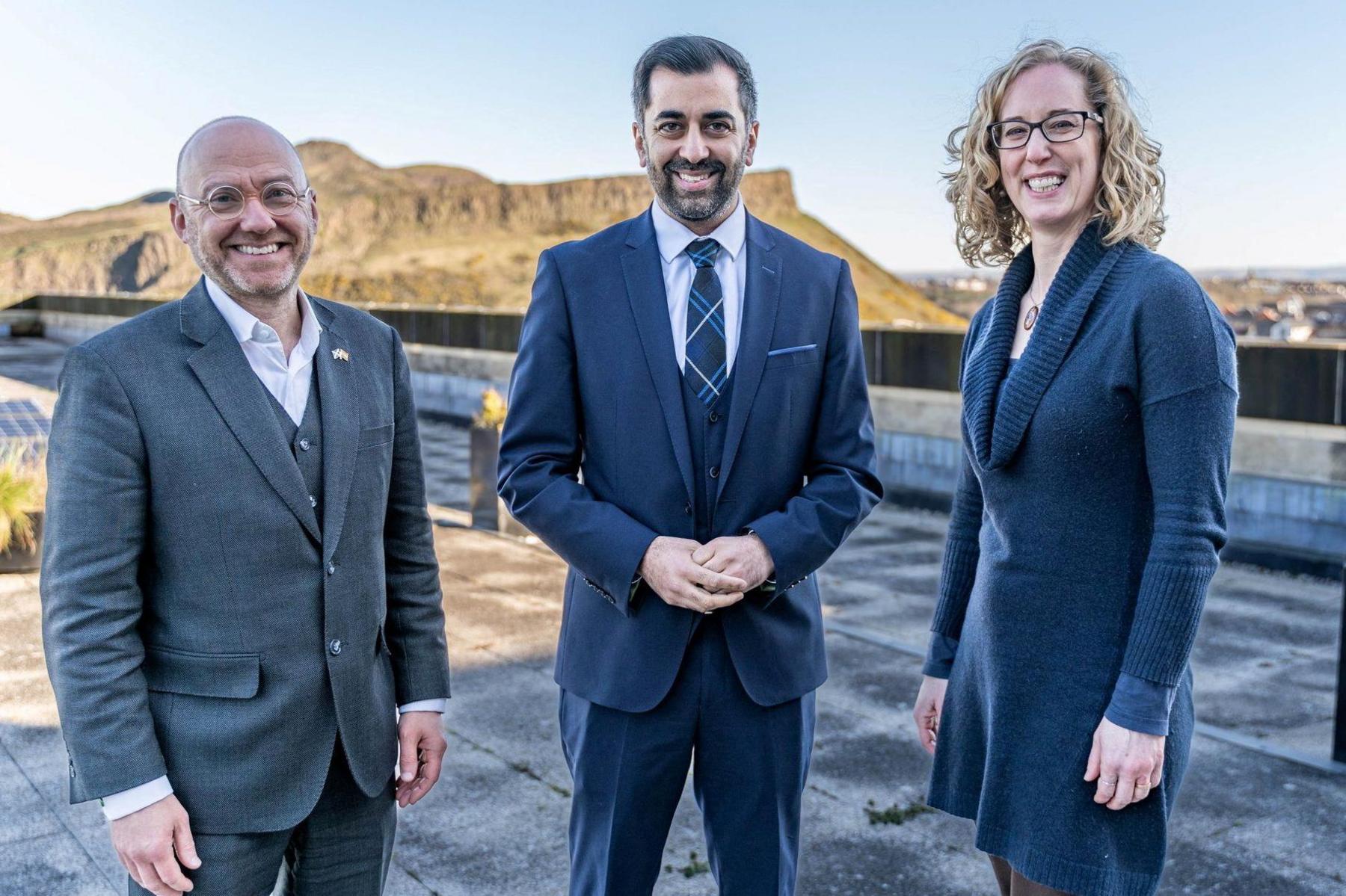 A bearded man in a blue suit is flanked by a bald man in a grey suit and a blond haired woman in a blue dress