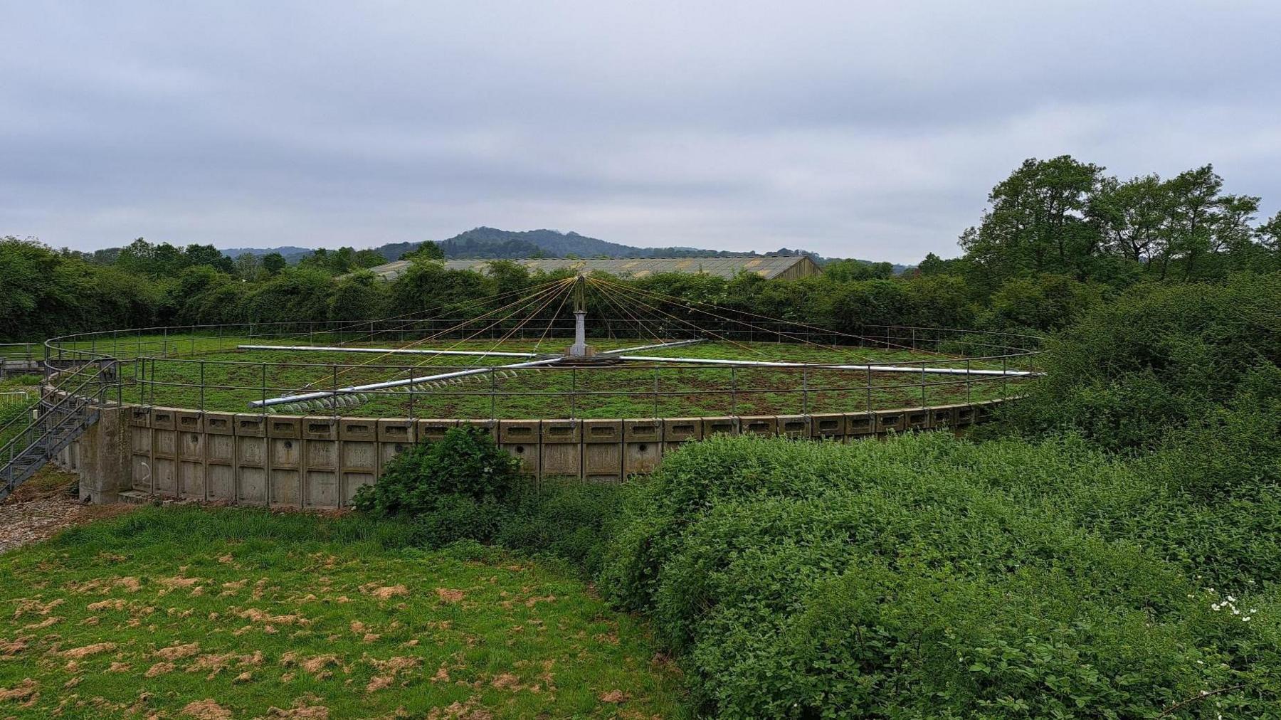  A large circular water treatment plant sits in the middle of a field. It resembles a huge wheel laying on its side, with metal railings extending from the centre out to the rim. On the surface of the plant appears to be mud and grass. 