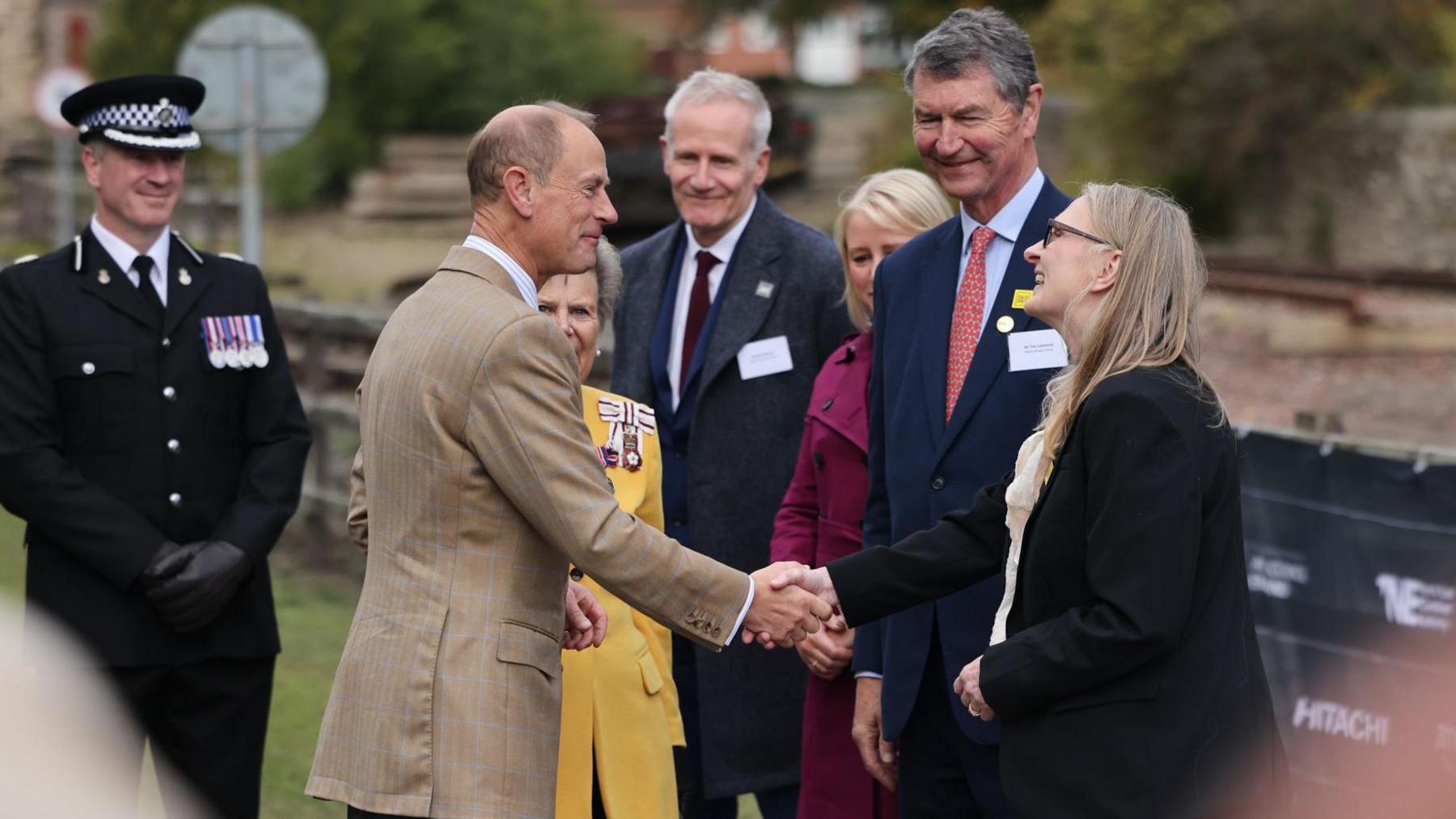 The Duke of Edinburgh, who is wearing a tan suit and has short fair hair that is thinning on top smiles as he shakes hands with a woman with long blonde hair. Several other people stand round and watch